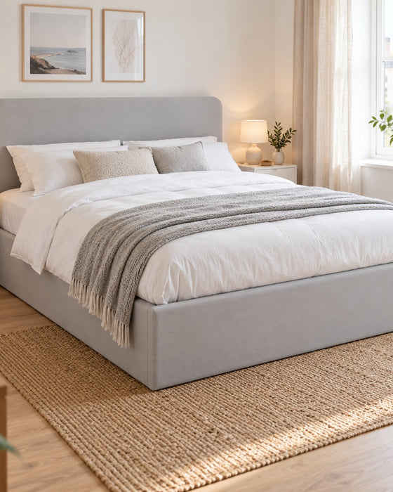 Brompton side lift ottoman bed in soft grey velvet with a rounded headboard, in a bright bedroom (AI-generated illustration of this frame; bedding for illustration, mattress not included)