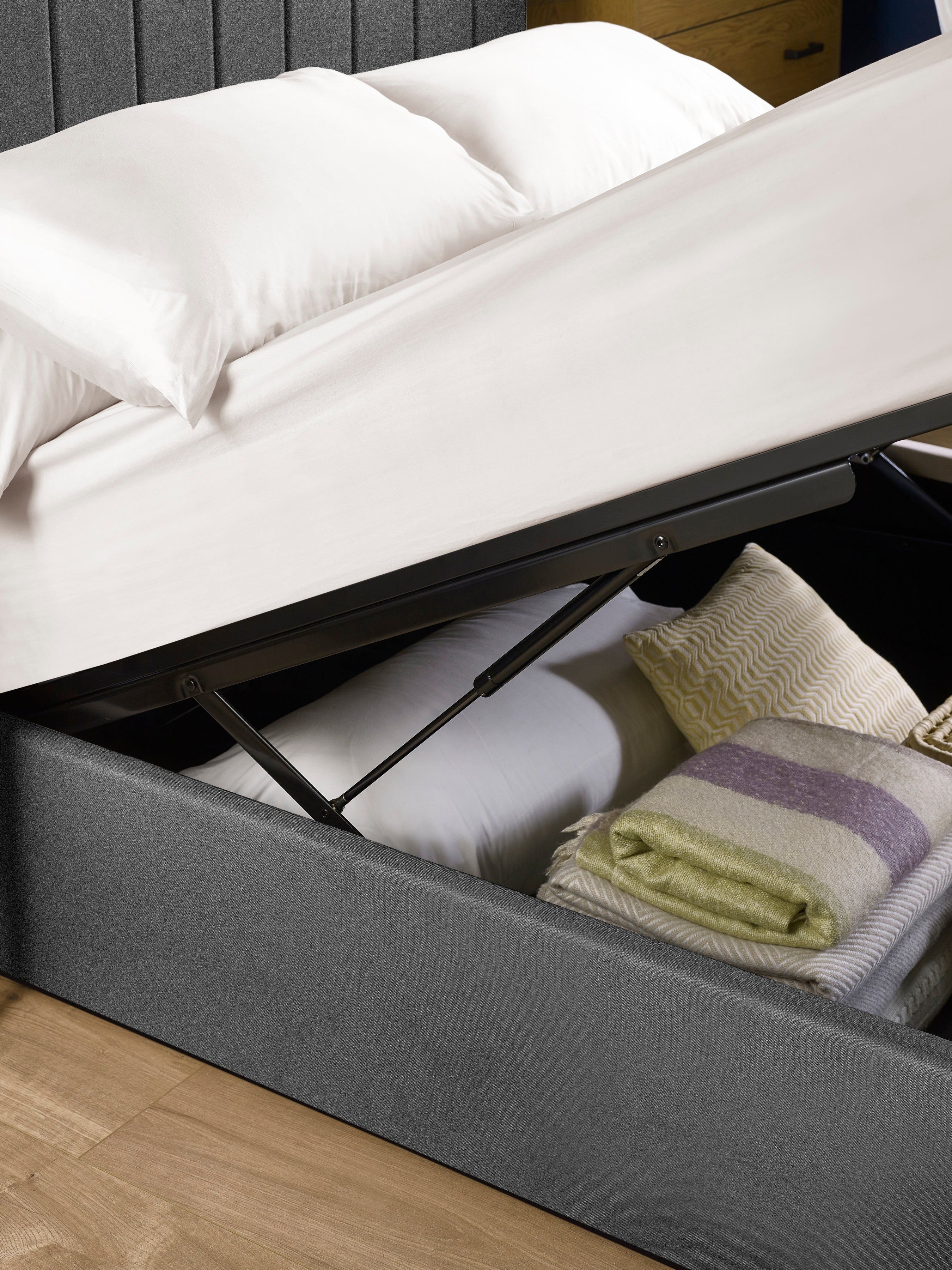 Aylen ottoman bed: gas lift raising the dark grey base over folded blankets, close up
