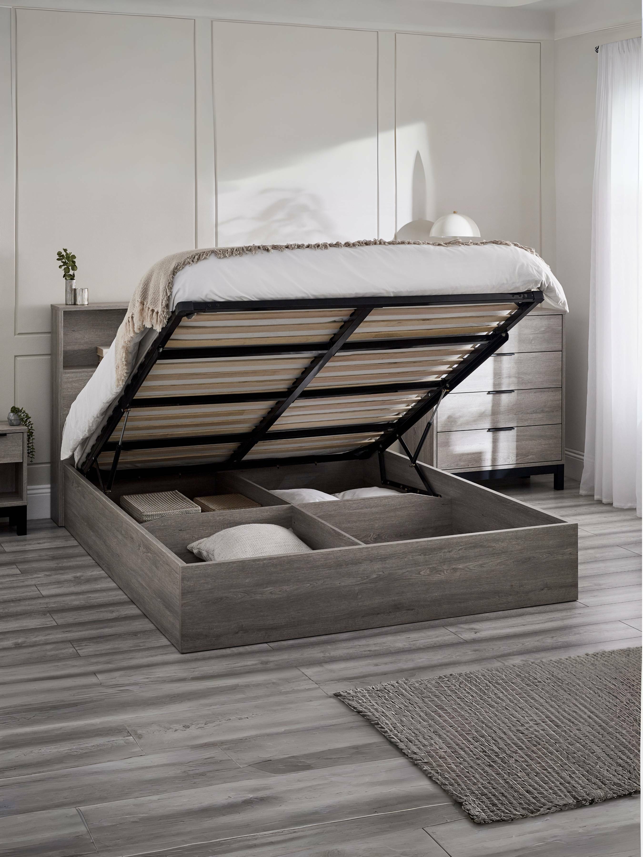 Bali grey oak effect ottoman bed with the gas lift base raised over the storage, in a bedroom