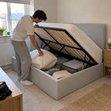 Man lifting the Brompton ottoman base from the side to put away a winter duvet (AI-generated illustration of this frame; bedding for illustration, mattress not included)